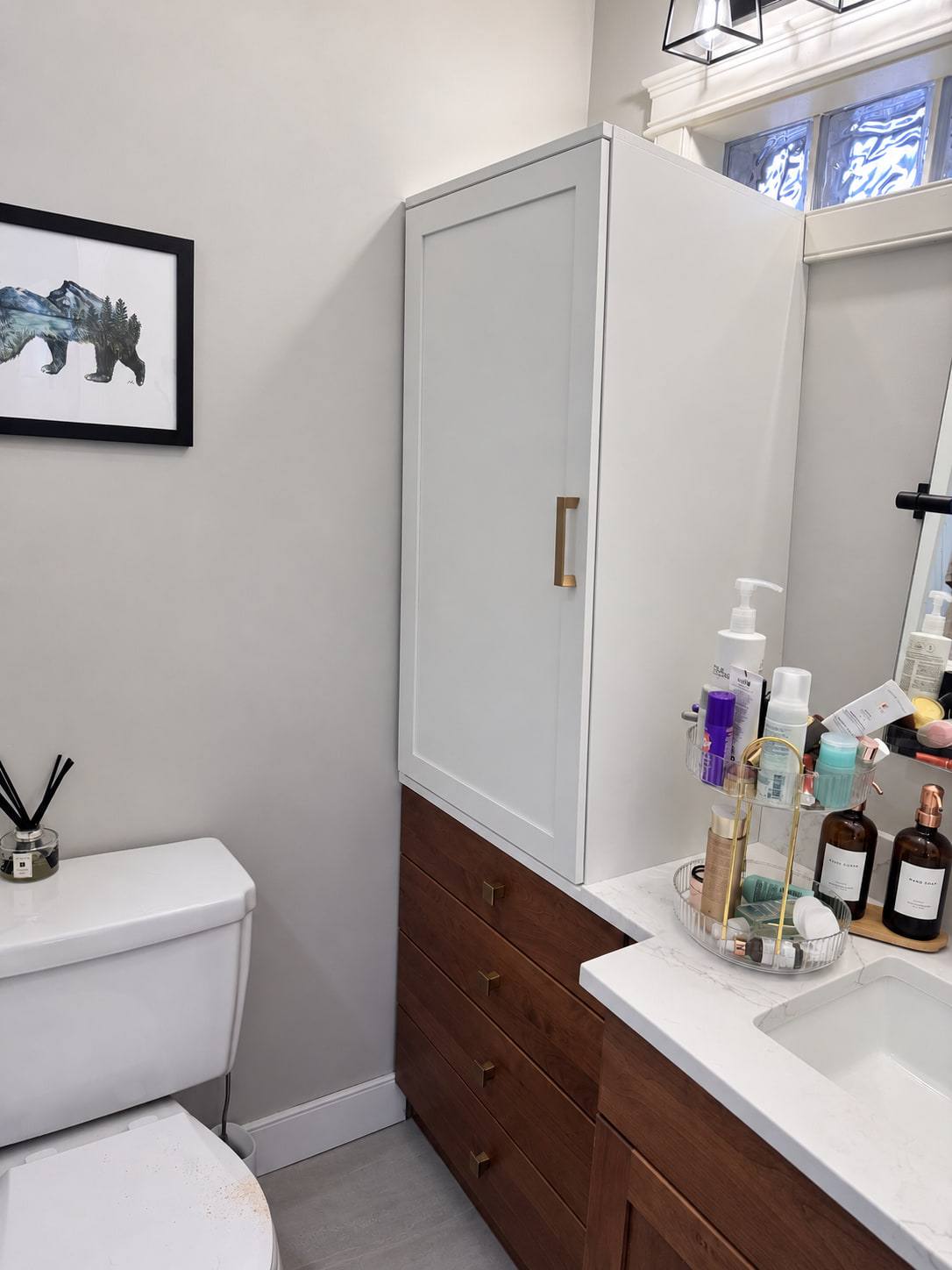 Bathroom vanity with wood drawers and a white top beside a tall white linen cabinet with a brass pull